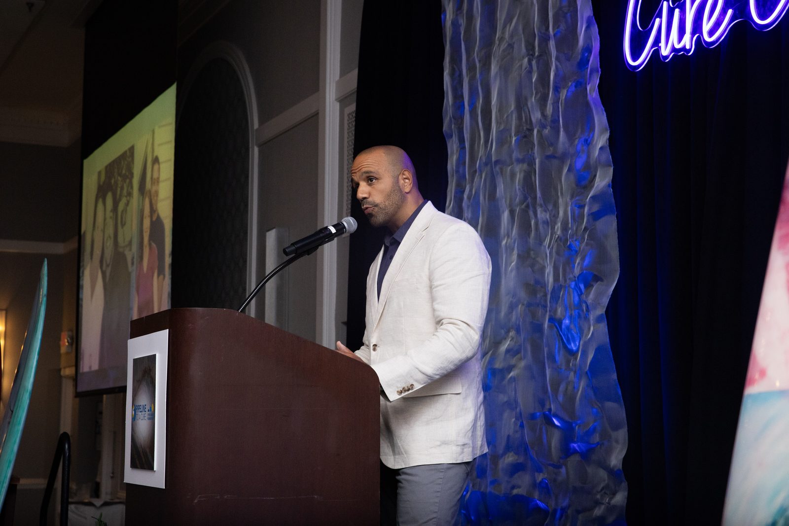 Ray speaking at a podium at the Pipeline to a Cure event, with blue stage lighting.