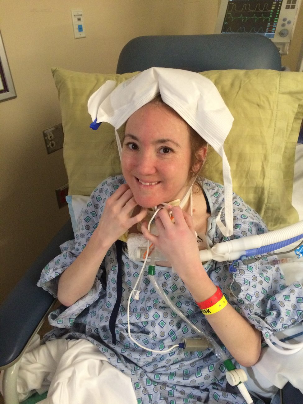 Rebecca smiling in a hospital chair with a white face mask resting on top of her head like a hat.