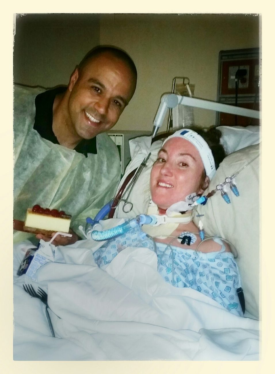 Ray in a protective gown holding a slice of cheesecake next to Rebecca, who smiles in a hospital bed.