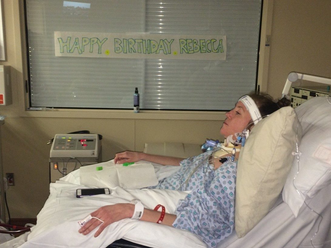 Rebecca resting in a hospital bed in front of a window with a handmade Happy Birthday Rebecca banner.