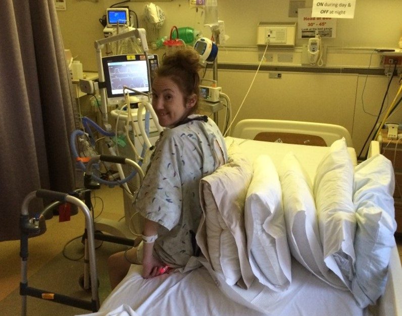Rebecca sitting on the edge of a hospital bed, looking back at the camera with a grin, with a walker and ventilator beside her.
