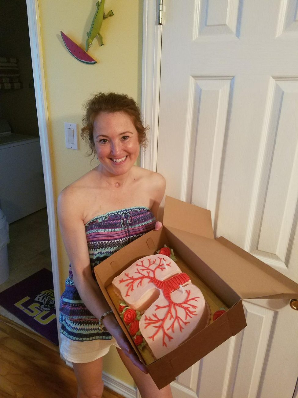 Rebecca smiling in a kitchen doorway, holding a box with a lung-shaped cake decorated with red airways.