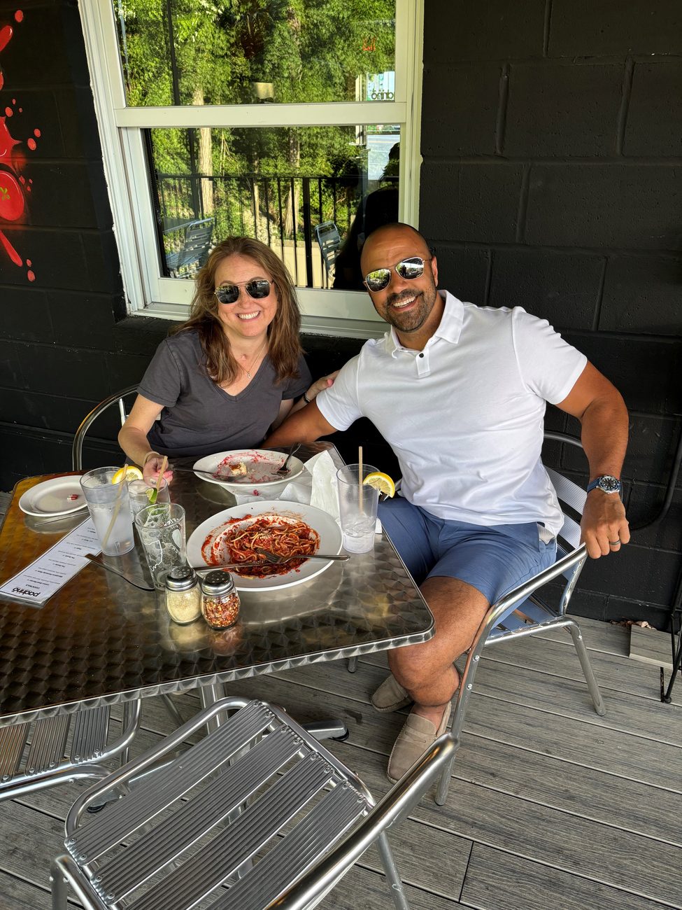 Ray and Rebecca in sunglasses at an outdoor table with plates of pasta, smiling at the camera.