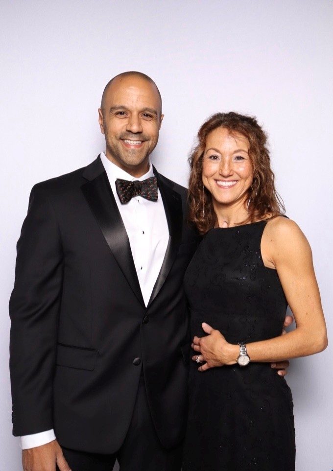Ray in a tuxedo and bow tie and Rebecca in a black dress, smiling together against a white backdrop.